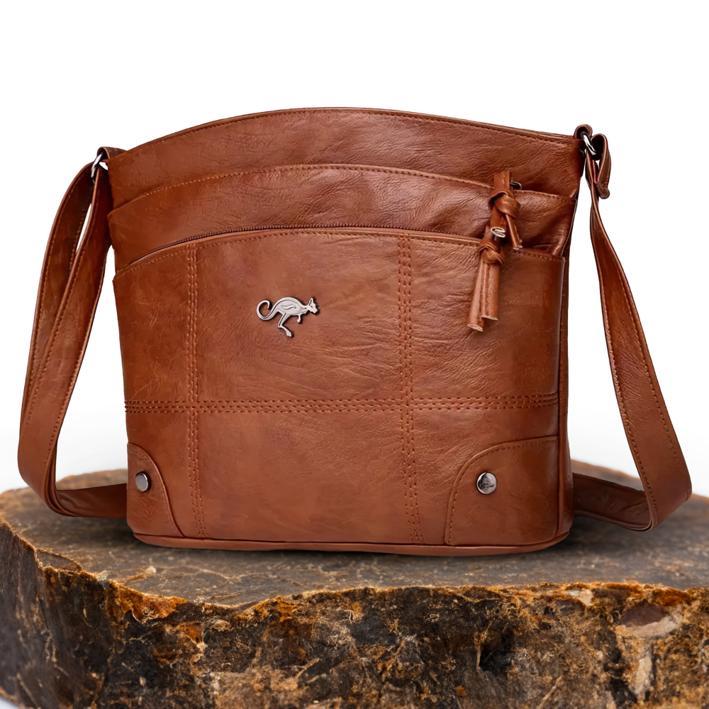 A brown leather shoulder bag with a curved silhouette, external pockets, and a secure zip closure, displayed on a natural stone surface.